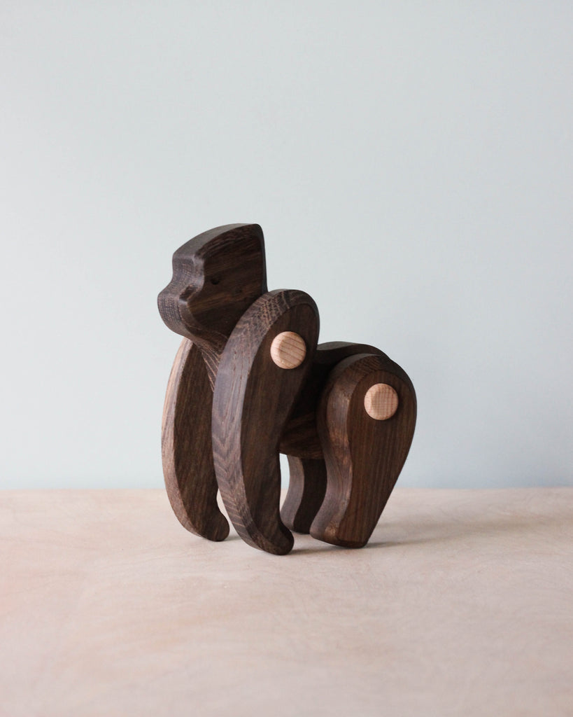 The Handmade Wooden Gorilla features a smooth, minimalist design with prominent wood grain and lighter circular joints, standing on a light surface against a plain pale blue background.