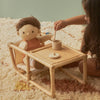 A child plays with a Wooden Doll Feeding Set seated at a small pinewood table, pretending to pour tea from a tiny pot into a cup, on a plush cream-colored rug against a pale green background.
