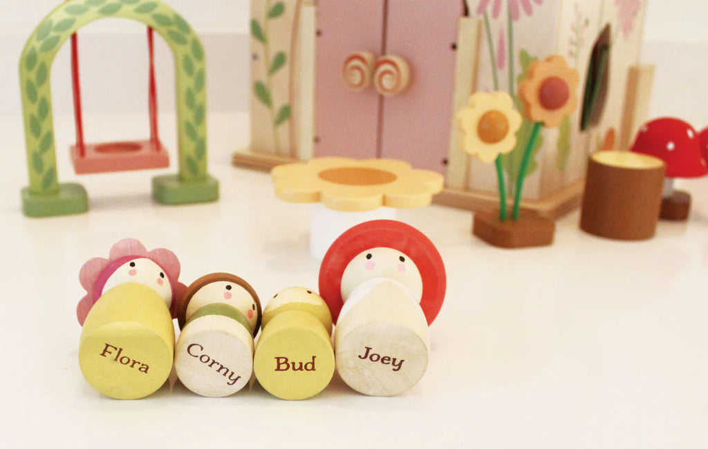 The Rosewood Cottage set charmingly displays four wooden peg dolls with flower-themed designs—Flora, Corny, Bud, and Joey—in front of a woodland cottage. The enchanting scene includes a toy house with a swing and delicate floral accents, evoking the magic of an imaginative meadow.