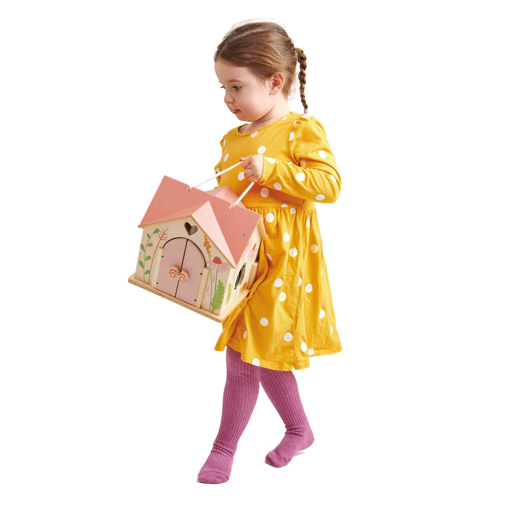 A young girl in a yellow polka dot dress and pink tights carries the Rosewood Cottage toy birdhouse with a pink roof and decorative plants. She walks barefoot against a white background, echoing the charm of a magical woodland cottage.