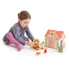 A young child with braided hair, in a floral dress and pink tights, is playing with the Rosewood Cottage toy set. It includes a house, archway, mushroom, and flower pieces on a playmat resembling a magical meadow.