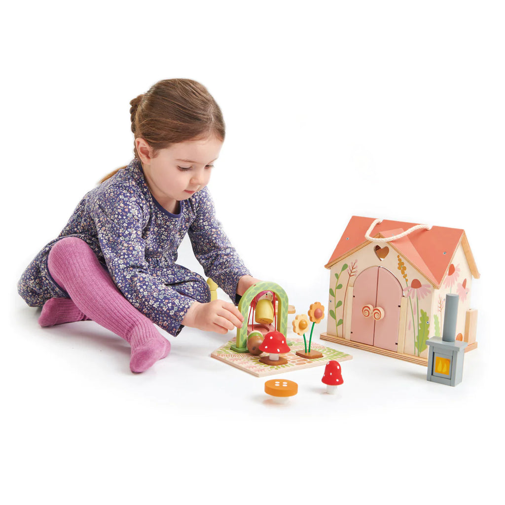 A young child with braided hair, in a floral dress and pink tights, is playing with the Rosewood Cottage toy set. It includes a house, archway, mushroom, and flower pieces on a playmat resembling a magical meadow.