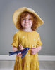A young girl with curly red hair wearing a yellow dress and a straw hat, holding a blue toy airplane with "Swifty Bird" on it, standing in front of a gray background