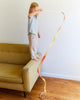 A child with long hair stands barefoot on the arm of a mustard-yellow couch, engaging in open-ended play. The child is holding Sarah's Silk Boho Streamer Wand, which flows downwards. The room features a white wall and wooden floor.