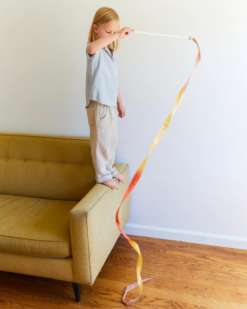 A child with long hair stands barefoot on the arm of a mustard-yellow couch, engaging in open-ended play. The child is holding Sarah's Silk Boho Streamer Wand, which flows downwards. The room features a white wall and wooden floor.