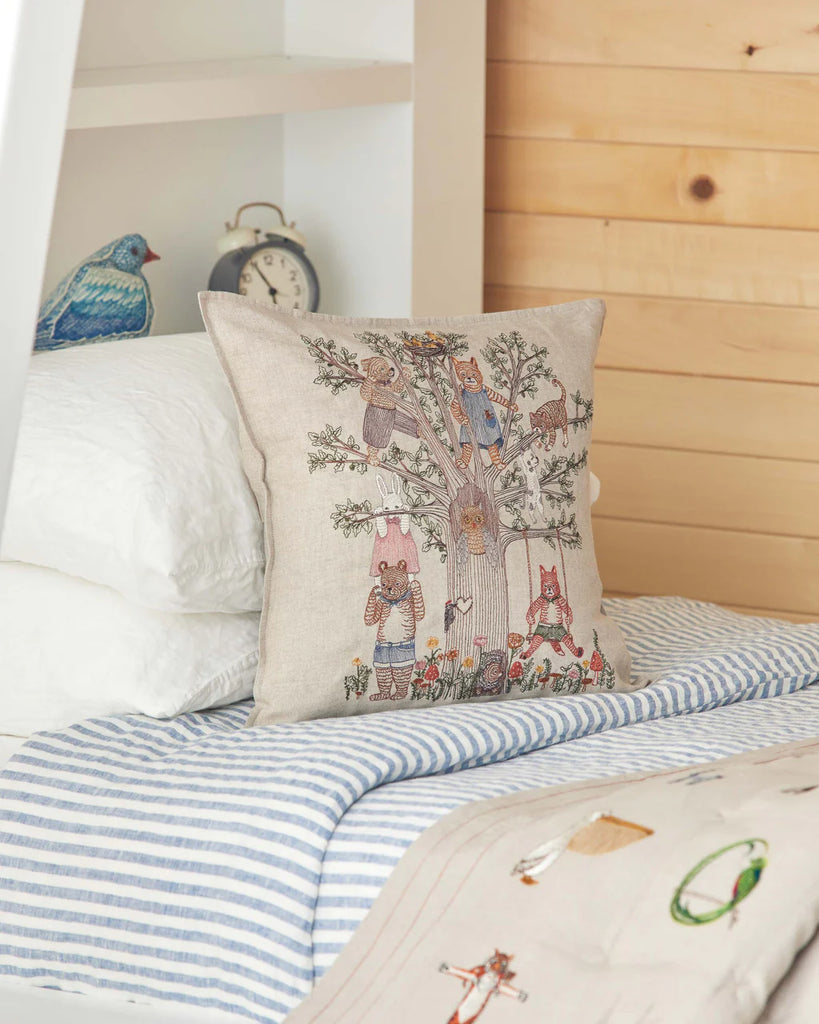 Throw pillow with woodland animal design, featuring creatures in clothes around a tree, on a bed with striped sheets, next to a white pillow and a Coral & Tusk Tree of Fun pillow, in a wooden cabin setting.