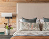 A cozy bedroom featuring a beige upholstered headboard, pocket pillows with woodland friends like the Coral & Tusk Hikers Pillow, a brown and white blanket, and a wall sconce. A small white vase with flowers and a book sit on the wooden nightstand against rustic wood walls for Camp-inspired decor.