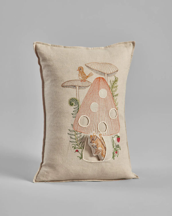 Decorative pillow featuring the Coral & Tusk Mushroom House Pocket Pillow, with large polka dots, surrounded by small plants and a foraging chipmunk, on a neutral fabric background.