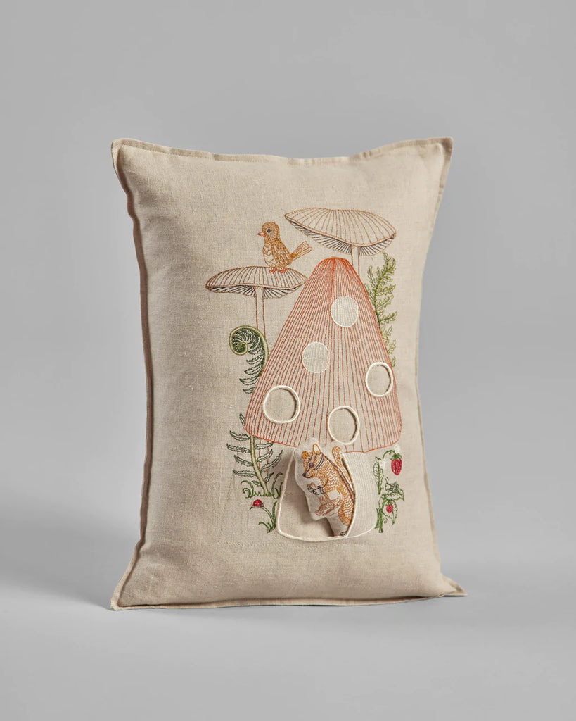 Decorative pillow featuring the Coral & Tusk Mushroom House Pocket Pillow, with large polka dots, surrounded by small plants and a foraging chipmunk, on a neutral fabric background.