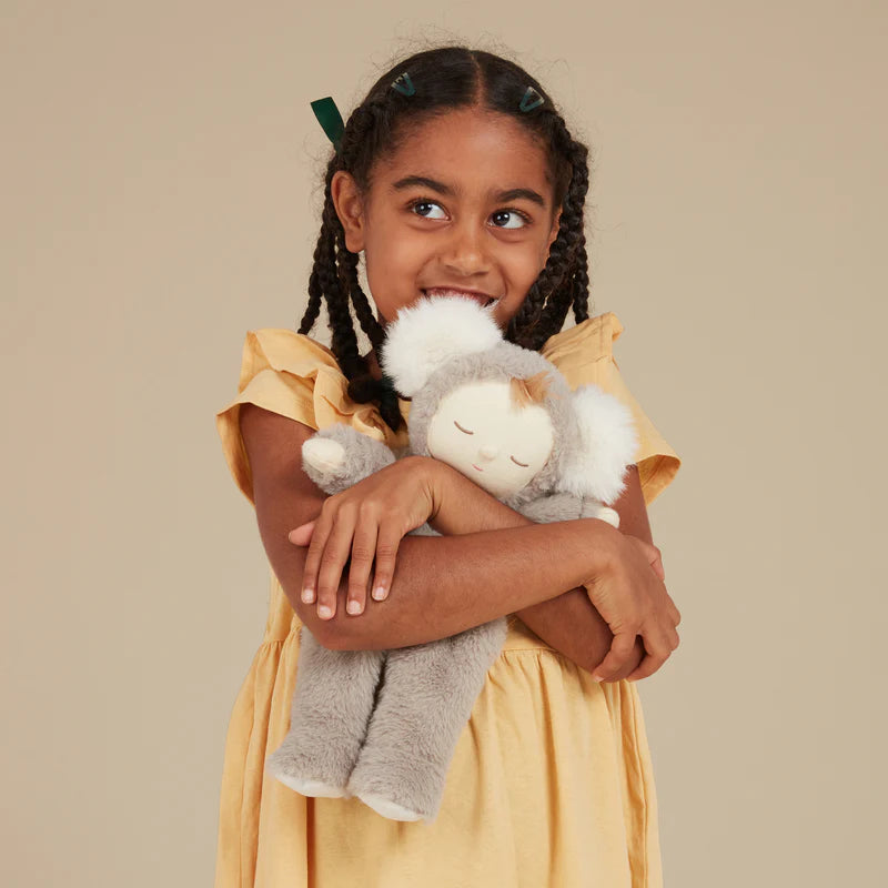A young girl with braided hair and green hair clips, wearing a yellow dress, smiling and hugging an Olli Ella Koala Moppet Doll against a neutral background.
