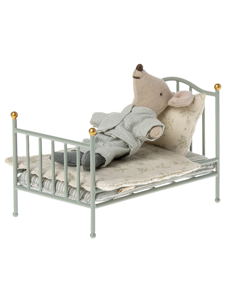 A small toy bear draped with a gray knitted blanket, lying on a Maileg Miniature Bed with gold accents, placed against a plain white background.