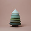 A minimalist, handmade wooden tree figurine with seven stacked, differently colored layers transitions from white at the top to dark green at the bottom, set against a light brown background. Crafted using non-toxic paint, it exemplifies sustainable responsibility. This is the Raduga Grez | Fir Tree Stacker.