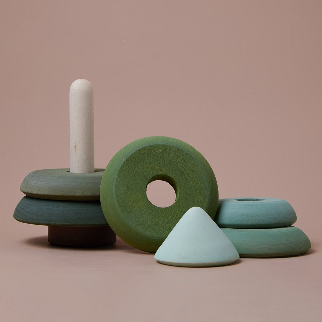 A Raduga Grez | Fir Tree Stacker with rings and a cone in pastel green and blue hues sits on a light brown surface against a matching backdrop. Painted with non-toxic paint, the toy is partially disassembled, with some rings stacked on the peg and others placed beside it.