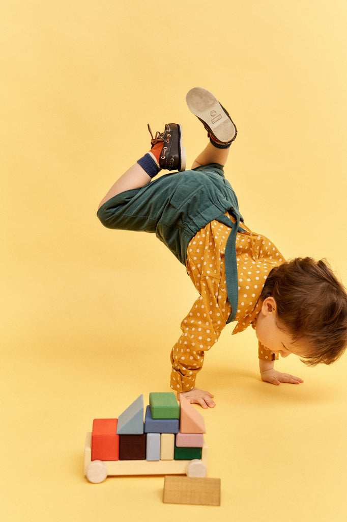 A young child in a yellow polka-dot shirt and green shorts playfully balances on one hand against a vivid yellow background. Nearby, a small cart holds Raduga Grez Vehicle Blocks finished with eco-friendly paint, adding a splash of creativity to this joyful scene.