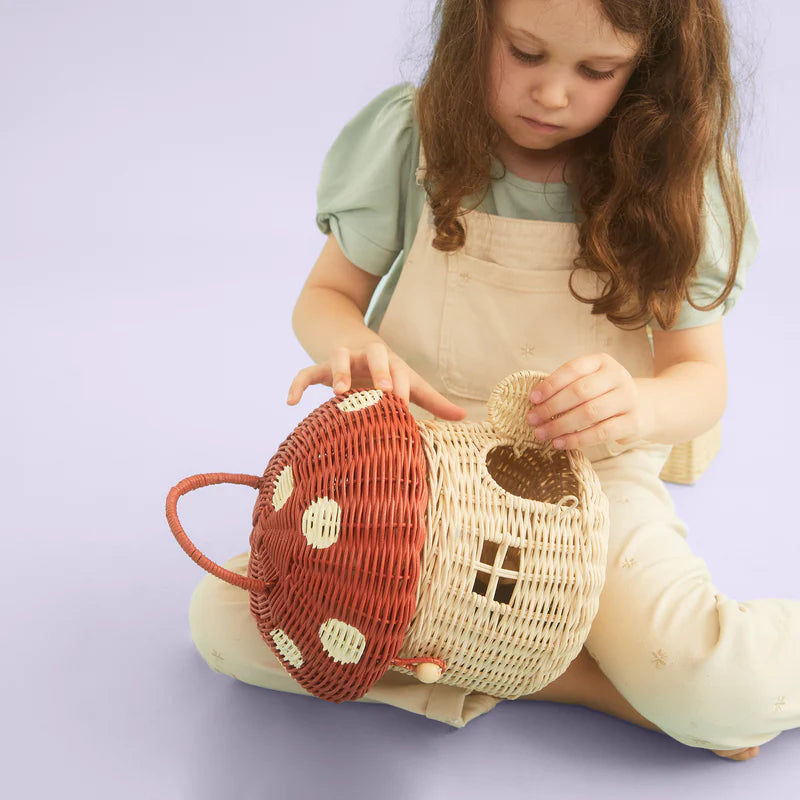 A young child examines a Olli Ella Red Mushroom Basket, woven in red and white, sitting on a light purple background.