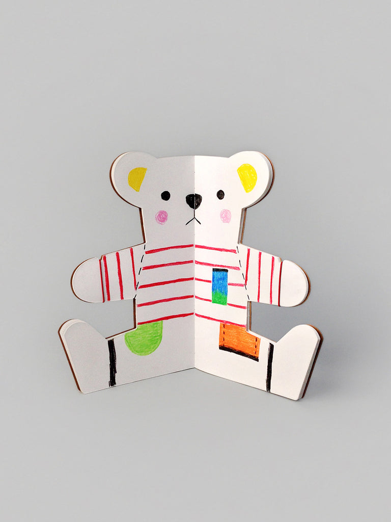 A colorful paper craft of a teddy bear with a striped red and white shirt, featuring multicolored patches on the body, arms, and ears, set against a plain gray background from The Teddy Bear Drawing Book.