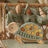 A cozy wicker crib is adorned with a hanging, double-decked holder filled with wicker and wooden toys. In the foreground, a children's night light shaped like a Little Lights Turtle Lamp emits a gentle glow from its cut-out patterns, resting on a decorative cushion with a leafy design.