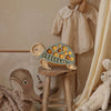 A cozy nursery scene features a wooden stool with a cute Little Lights Turtle Lamp on top, casting a gentle glow. The wall showcases a charming illustration of a whale, while a baby outfit hangs neatly nearby. A fluffy white teddy bear sits close by, adding to the room's warm and welcoming atmosphere.