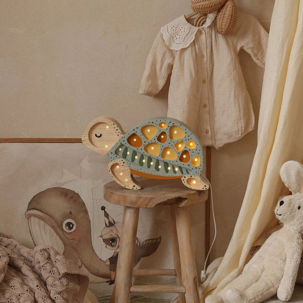 A cozy nursery scene features a wooden stool with a cute Little Lights Turtle Lamp on top, casting a gentle glow. The wall showcases a charming illustration of a whale, while a baby outfit hangs neatly nearby. A fluffy white teddy bear sits close by, adding to the room's warm and welcoming atmosphere.