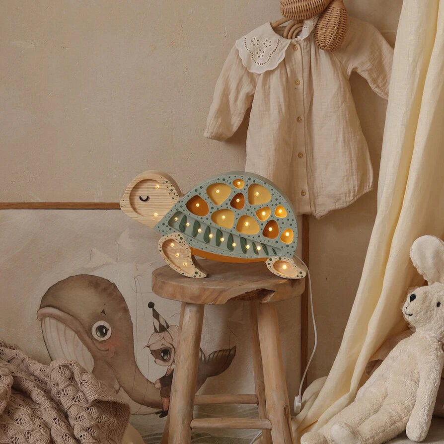 A cozy nursery scene features a wooden stool with a cute Little Lights Turtle Lamp on top, casting a gentle glow. The wall showcases a charming illustration of a whale, while a baby outfit hangs neatly nearby. A fluffy white teddy bear sits close by, adding to the room's warm and welcoming atmosphere.