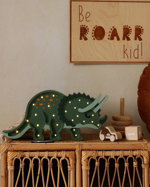 The Little Lights Triceratops Lamp, with its green dinosaur design and lit-up holes, stands proudly on a wicker shelf. Above it, a cheerful wooden sign proclaims, "Be ROARR kid!" Nearby, a stacking toy and small box enhance the nursery decor, while a round cushion softly complements the space.