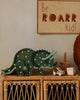 The Little Lights Triceratops Lamp, with its green dinosaur design and lit-up holes, stands proudly on a wicker shelf. Above it, a cheerful wooden sign proclaims, "Be ROARR kid!" Nearby, a stacking toy and small box enhance the nursery decor, while a round cushion softly complements the space.
