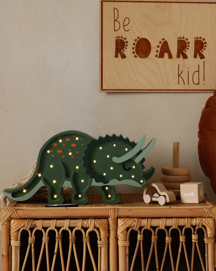The Little Lights Triceratops Lamp, with its green dinosaur design and lit-up holes, stands proudly on a wicker shelf. Above it, a cheerful wooden sign proclaims, "Be ROARR kid!" Nearby, a stacking toy and small box enhance the nursery decor, while a round cushion softly complements the space.