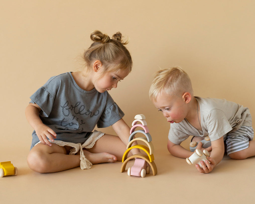 Two kids playing with  stacking toy with 7 pastel tone wooden arcs stacked into a rainbow shape. 