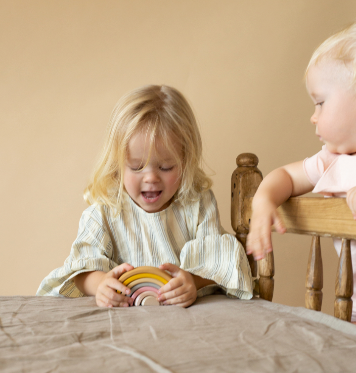 A kid playing with a stacking toy with 7 pastel tone wooden arcs stacked into a rainbow shape. 