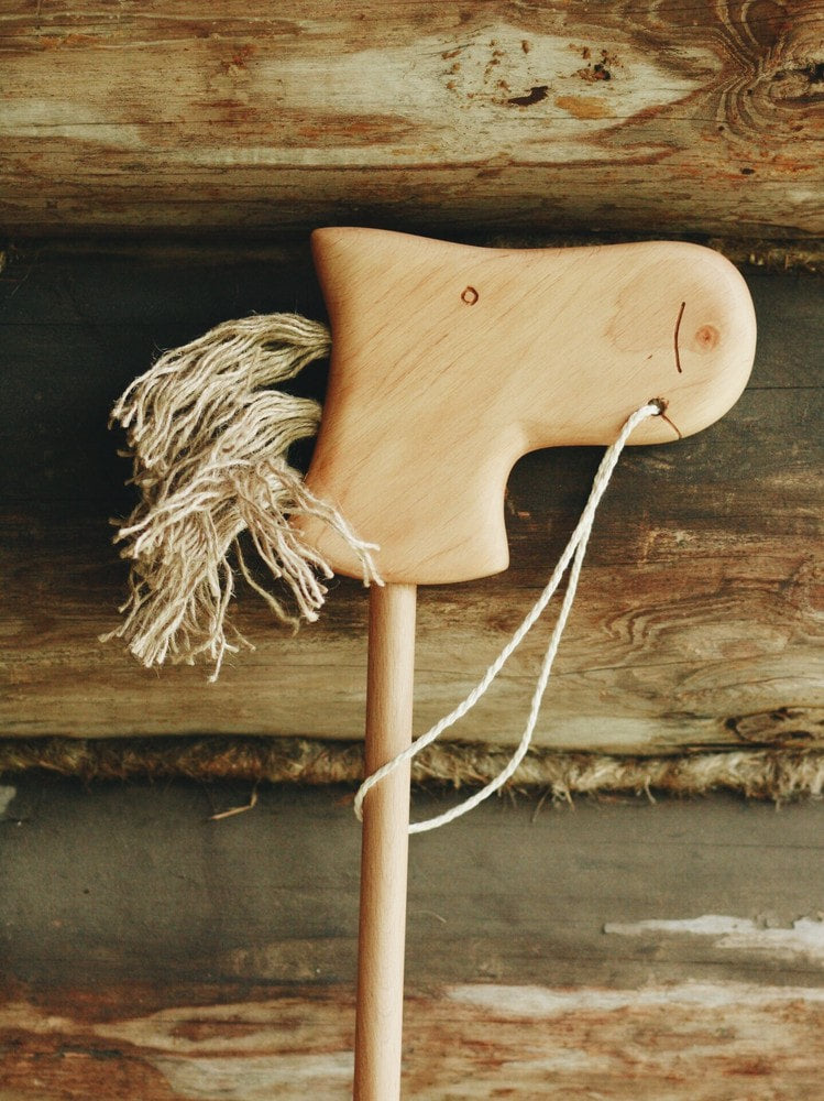 A Handmade Wooden Riding Horse, featuring a carved head and yarn mane, crafted from alder wood with string reins, leans against rustic log cabin-style planks.